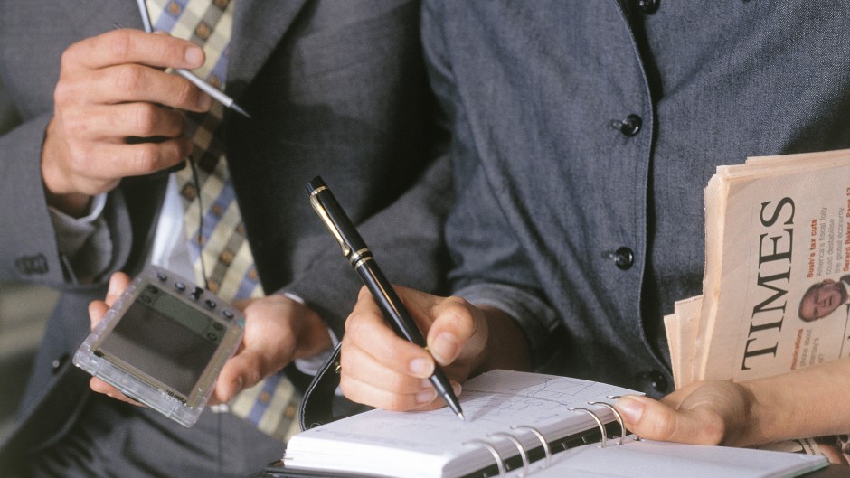 Close-up of hands of woman in suit writing on diary, hands of man in suit holding a palm-pilot at her side.