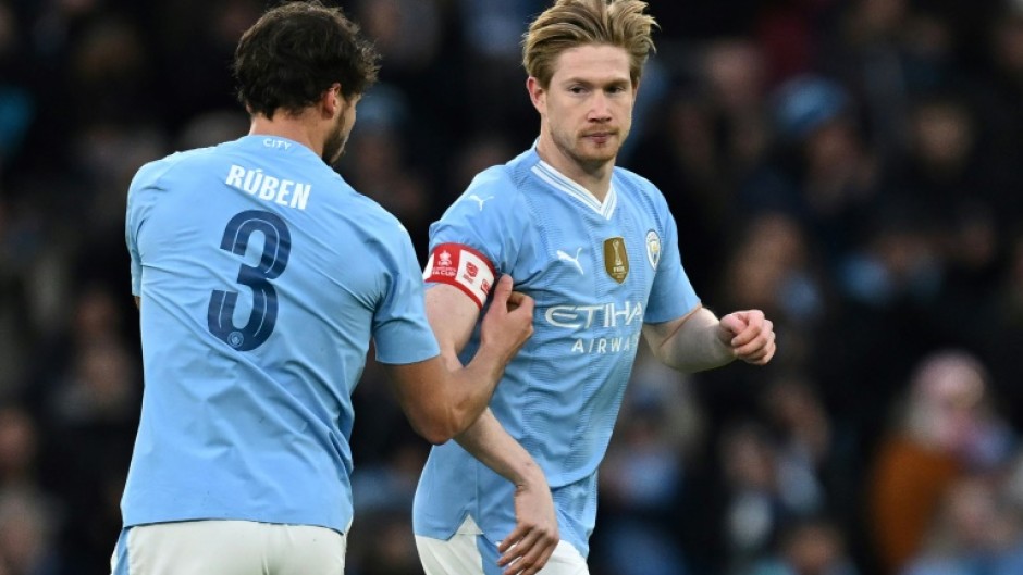 Manchester City's Ruben Dias (L) passes the captain's armband to Kevin De Bruyne