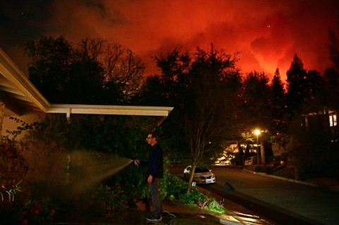 A man waters the front of his house as smoke and flames from the Palisades Fire burn toward the Encino neighborhood of Los Angeles, California