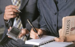 Close-up of hands of woman in suit writing on diary, hands of man in suit holding a palm-pilot at her side.